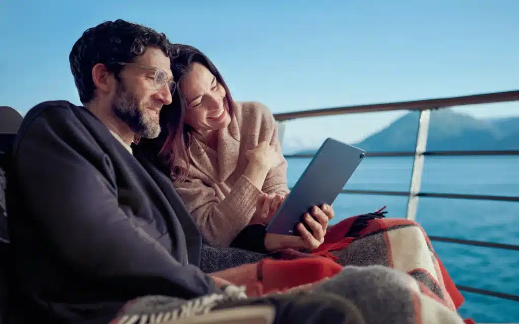 One male and one female Cunard passenger read from a device outside on an open deck.