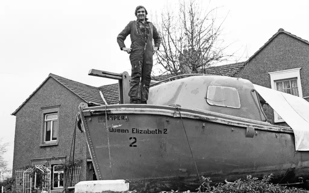 A male stands atop a QE2 life boat on dry land.