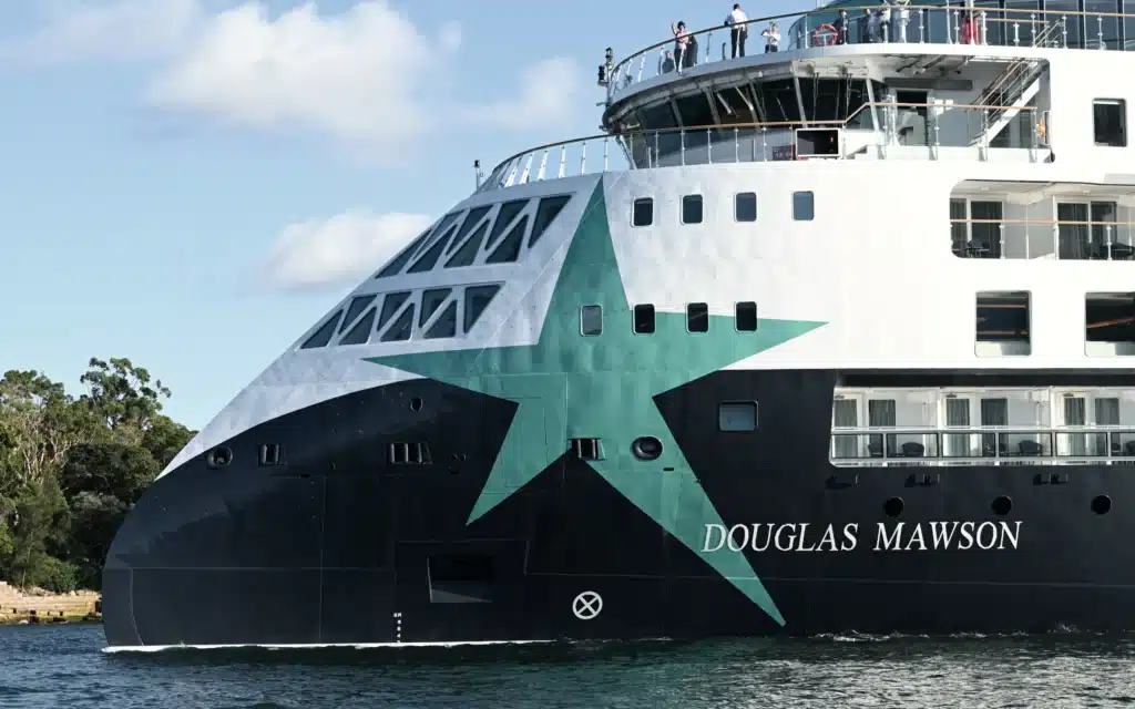 The bow of Aurora Expeditions' expedition ship Douglas Mawson, featuring a bold teal star on a dark hull, with passengers on the observation deck