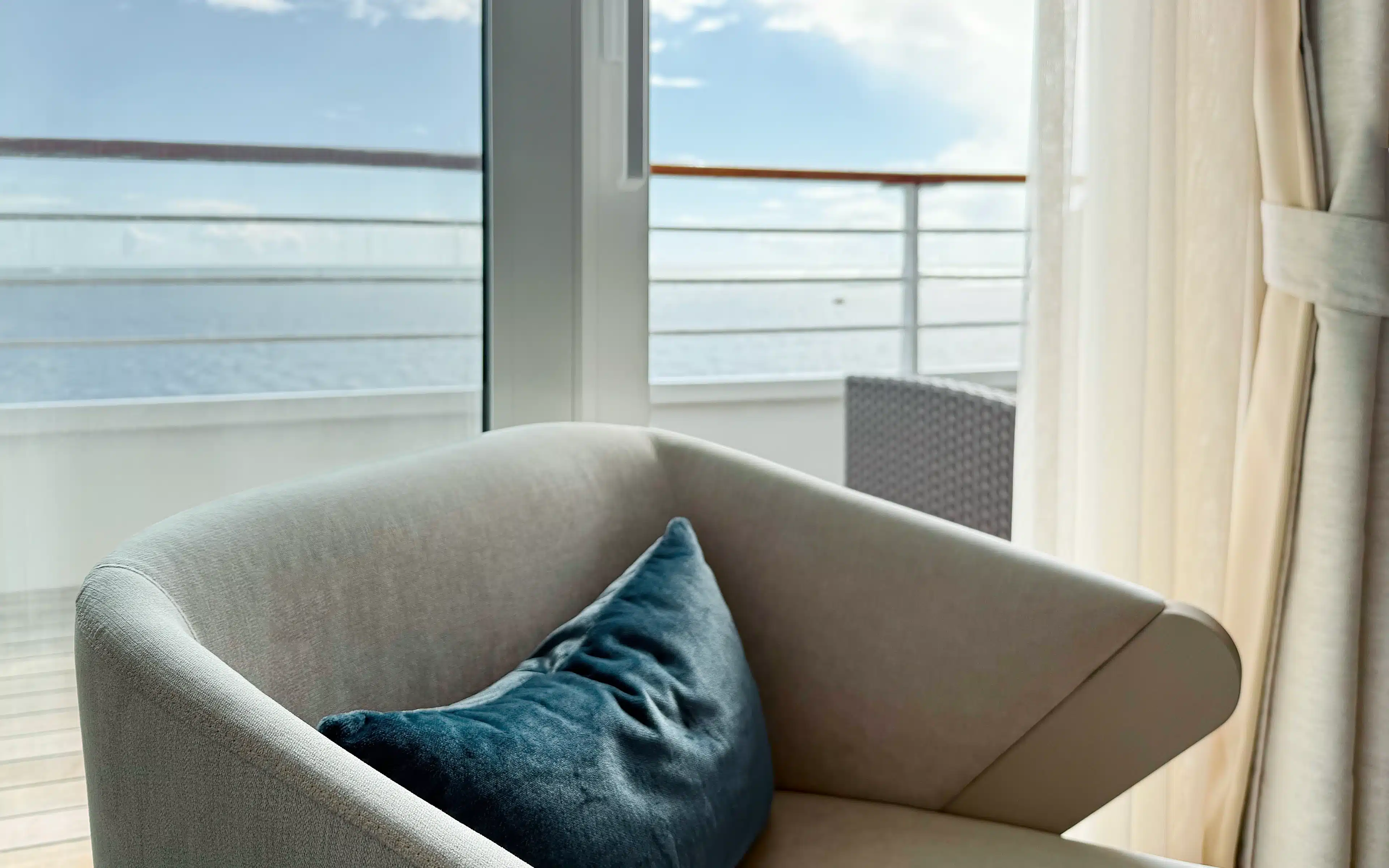 Looking out the doors of a Sapphire Veranda Suite toward the teak balcony, with an armchair in the foreground and blue ocean in the background.