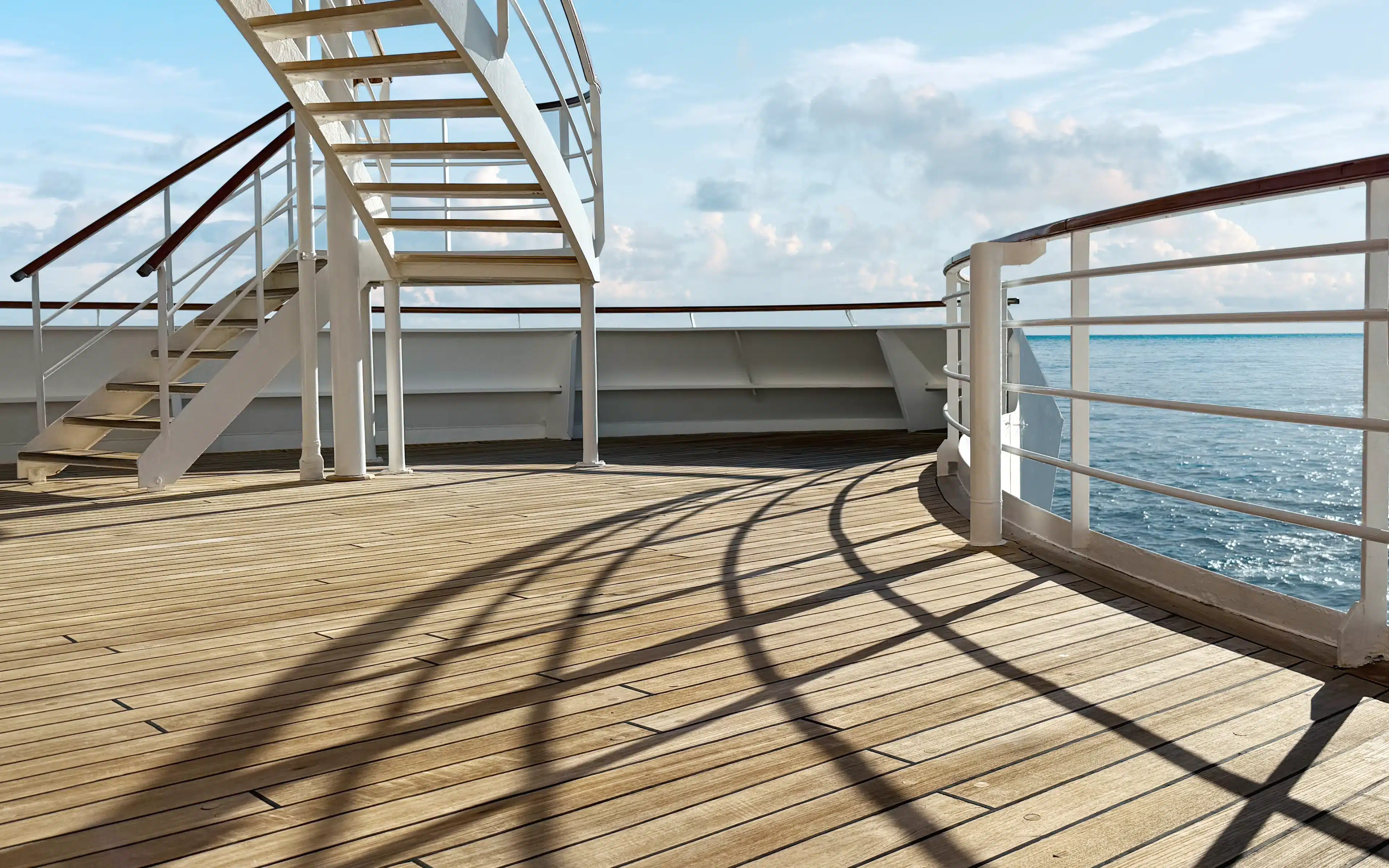 Spiral staircases and railings cast shadows on teak deck at aft of Crystal Serenity with ocean background.