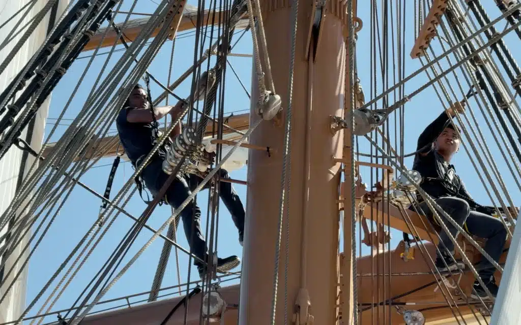 Two Sea Cloud Spirit crew members in safety harnesses working high in the rigging