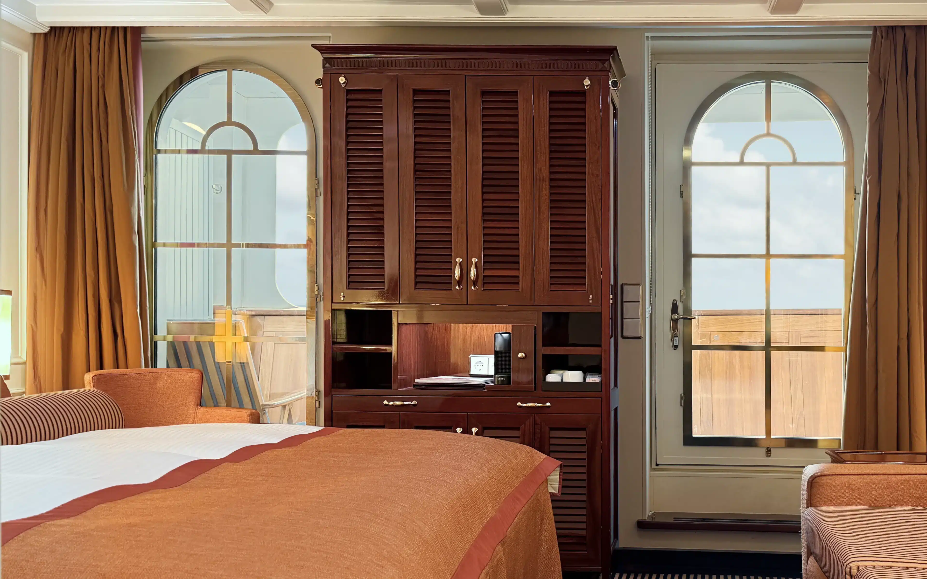 Sea Cloud Spirit Junior Veranda Suite showing the full room layout with terracotta king bed, mahogany storage unit, striped sofa, arched veranda door and windows, navy carpet and original sailing artwork