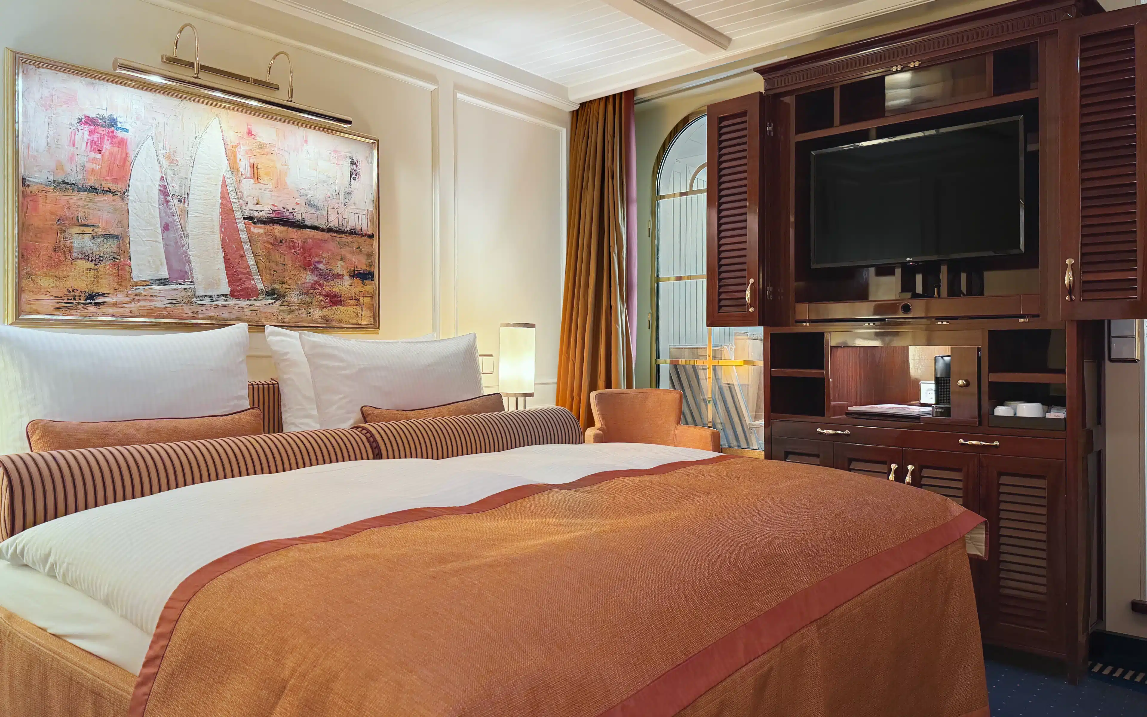 Sea Cloud Spirit Junior Veranda Suite with terracotta king bed, original sailing artwork above the headboard, mahogany entertainment unit with flat-screen television, arched veranda door and warm amber curtains