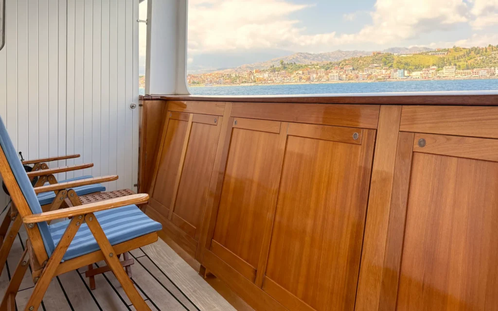 Sea Cloud Spirit Junior Veranda Suite private veranda with teak deck, timber paneling, striped blue cushion chairs and a Mediterranean coastal town in the background