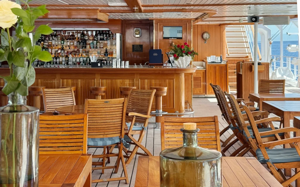 Sea Cloud Spirit Lido Bar by day with teak furniture, a fully stocked bar, red roses and the ship's rigging and open sea visible beyond