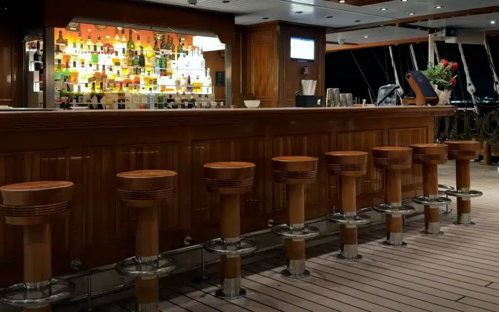 Sea Cloud Spirit Lido Bar at night with illuminated backlit spirits bottles, teak bar stools, mahogany counter and the darkened rigging visible beyond