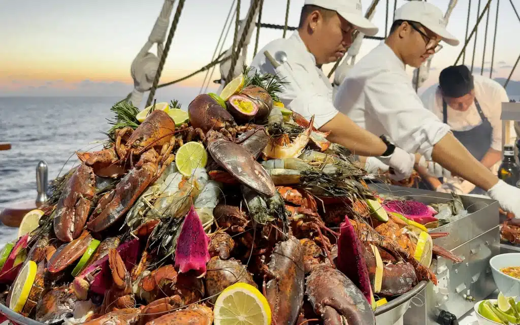 Sea Cloud Spirit deck barbecue lobster display piled high with whole lobsters, tiger prawns and dragon fruit, tended by chefs in whites at sunset
