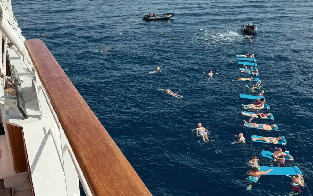 Guests swimming and floating on blue mats in the open ocean alongside Sea Cloud Spirit, with two safety tenders standing by