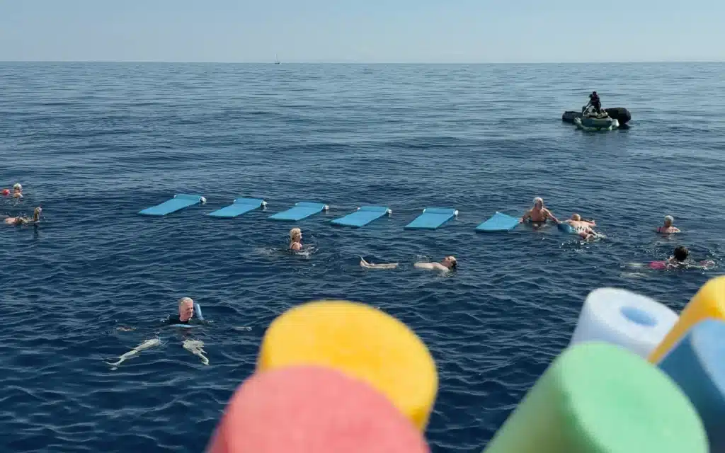 Wide view of Sea Cloud Spirit guests swimming in the open sea with floating mats and a safety tender, seen from the ship's swim platform