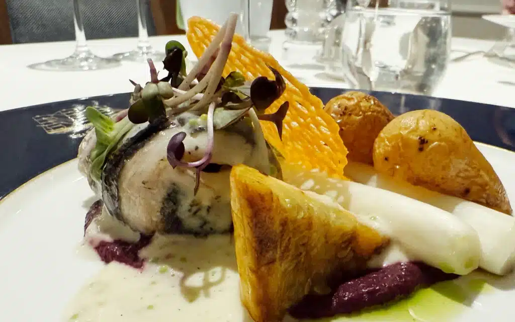 Sea Cloud Spirit restaurant sea bream fillet with white asparagus, purple potato purée, roasted new potatoes, pastry shard and herb cream sauce, on a navy rimmed plate