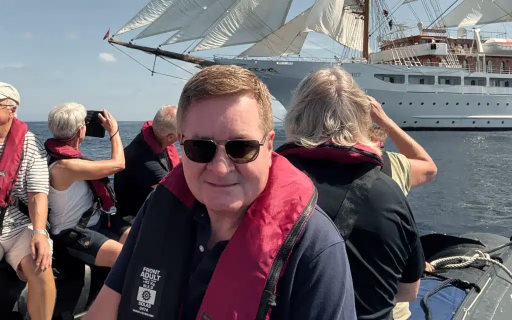 A guest in a life jacket aboard a Sea Cloud Spirit Zodiac tender, with the ship under sail visible in the background and fellow passengers photographing the scene