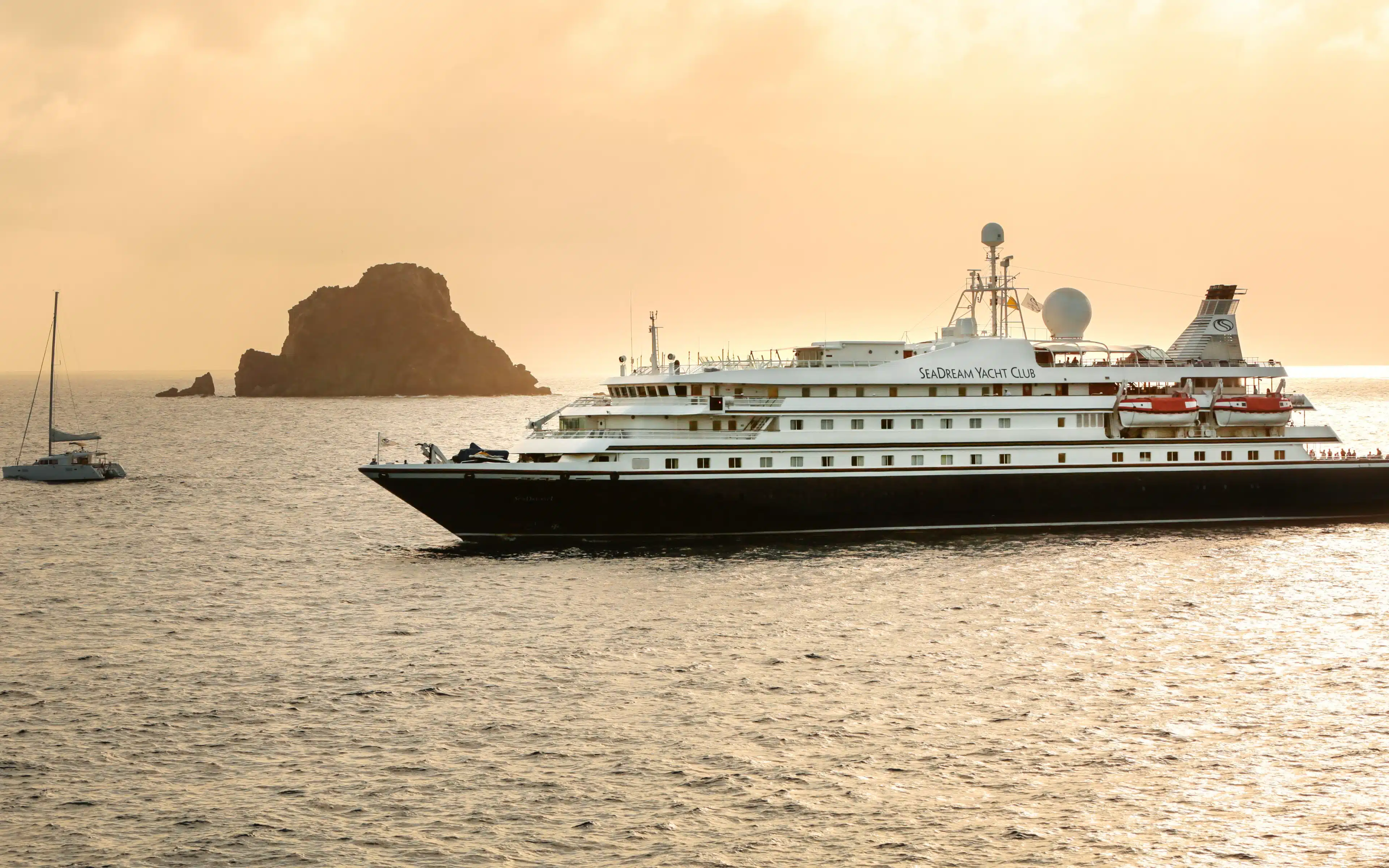 A SeaDream yacht at anchor during golden hour A SeaDream Yacht Club vessel anchored at dusk with a rocky islet silhouetted against a golden sky