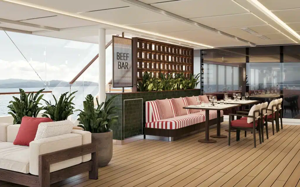 The outdoor deck dining area of Beef Bar on Crystal Grace, with teak timber decking, striped red and white sofa seating, a green tiled bar counter with tropical plants, and panoramic sea and mountain views