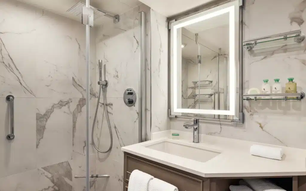 Marble bathroom in Oceania Marina's Concierge Veranda Stateroom, with a walk-in rainfall shower, illuminated vanity mirror, and Bvlgari toiletries on chrome shelving