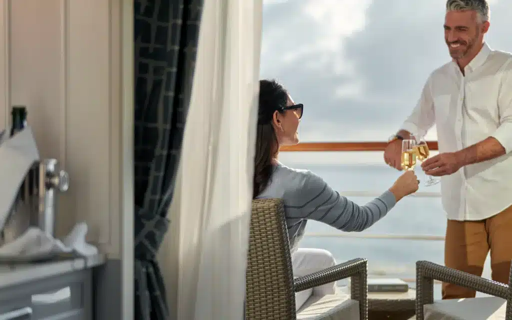 A couple toasting champagne flutes on the private veranda of an Oceania Marina Concierge Veranda Stateroom, with open ocean views beyond the railing
