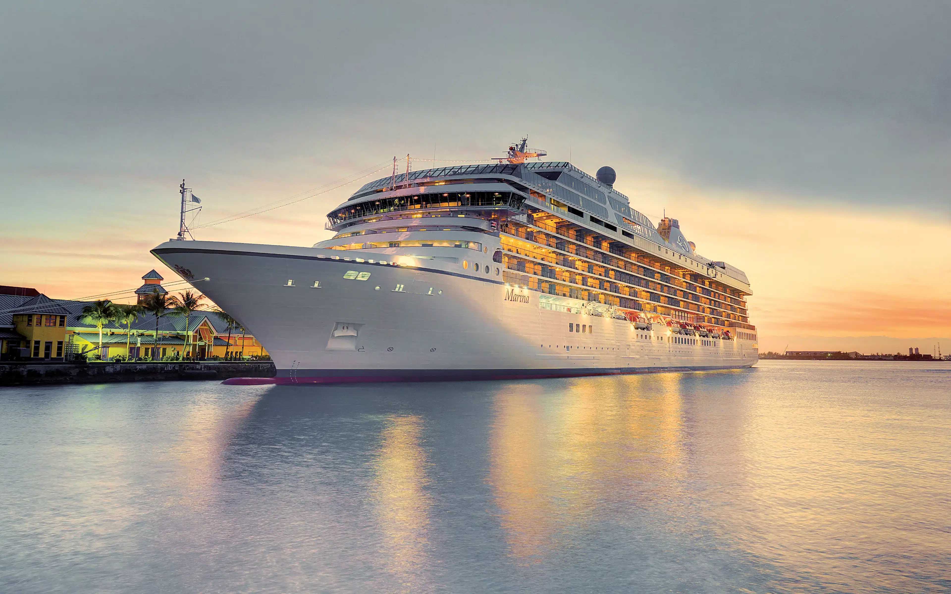 Oceania Marina cruise ship illuminated at dusk, docked at a tropical port with a pastel sunset sky reflected in calm water