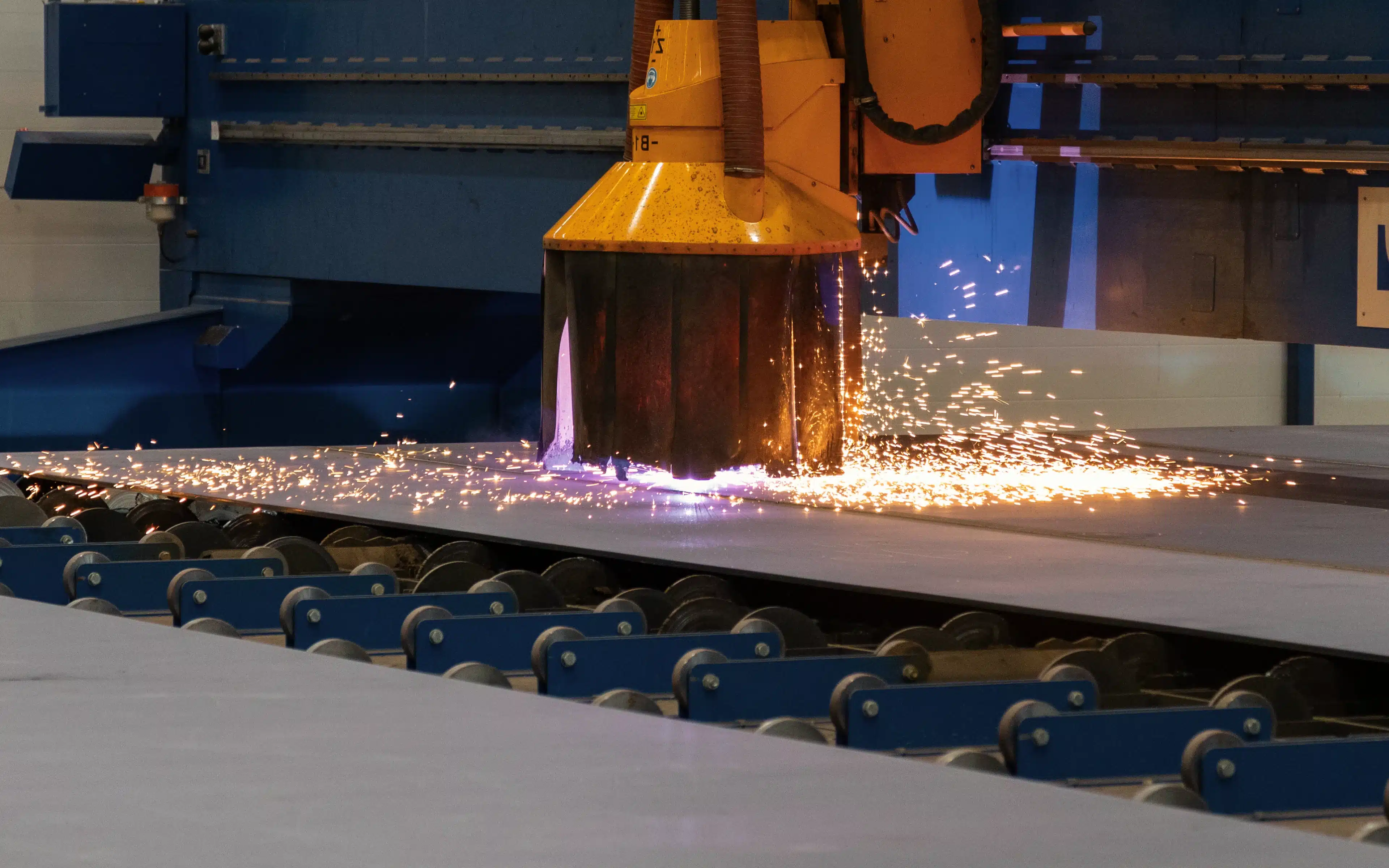An industrial plasma or laser cutting machine with an orange robotic arm cuts through a large steel plate on a blue roller conveyor, sending bright orange sparks and a violet flame across the surface in a shipyard manufacturing facility.