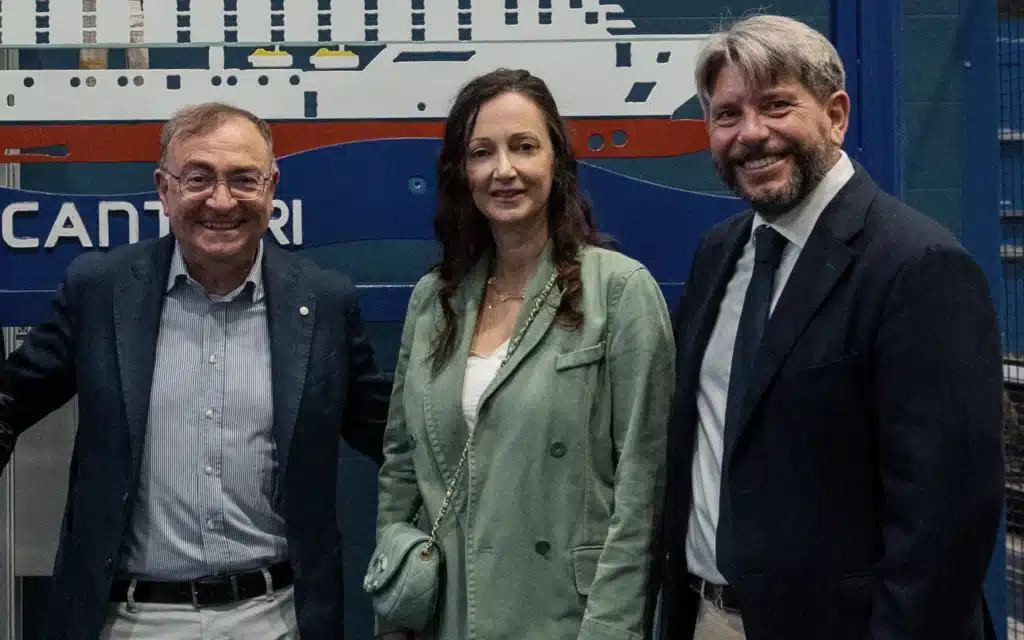Three smiling individuals—an older man in a navy blazer and striped shirt, a woman in a sage green double-breasted jacket, and a man in a dark navy suit with a tie—pose together in front of a large illustrated schematic of a cruise ship at a Fincantieri shipyard facility. Left to right: Luigi Matarazzo, Cristina Levis, and Bernie Leypold.