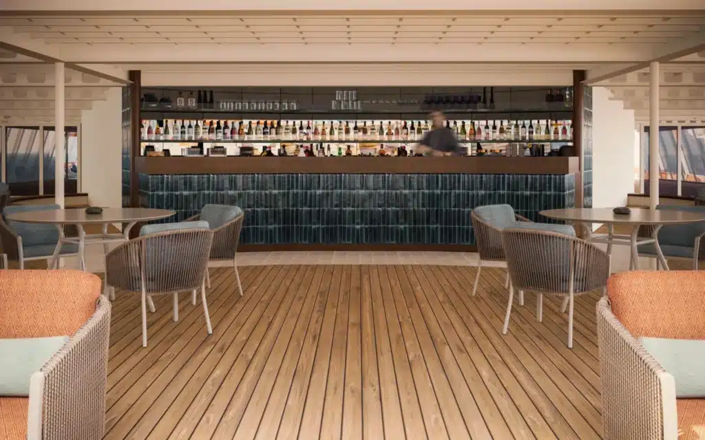 Crystal Serenity pool bar area following the 2026 drydock refit, featuring wide-plank teak decking, woven rope café chairs and round tables, coral-upholstered wicker sofas in the foreground, and a full-width bar with teal glazed ceramic tile facing, backlit shelving stocked with spirits bottles, and a bartender in motion behind the counter
