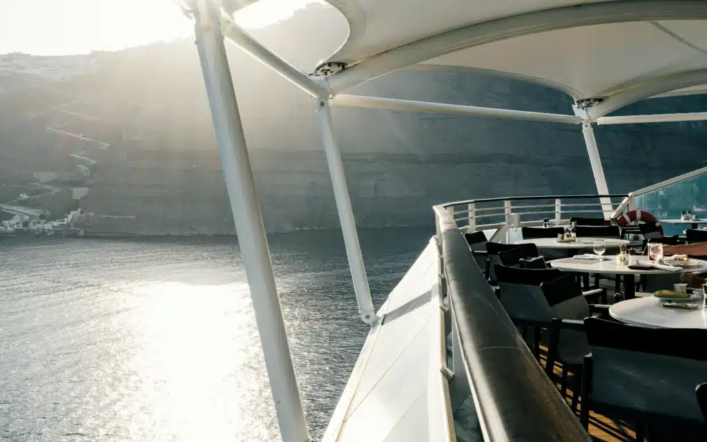 Canopied outdoor deck dining area aboard an Oceania Cruises ship, with dark chairs and round white tables set with stemware and dining plates, overlooking the dramatic volcanic caldera cliffs of Santorini, Greece, with sunlight scattering across the water below