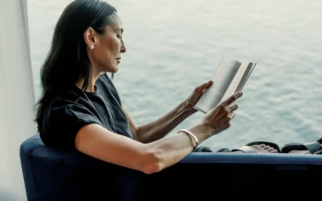 A dark-haired woman in profile wearing a black short-sleeve textured top, pearl drop earrings, and a rose gold bangle bracelet, reading an open paperback book while seated in a navy blue armchair beside a large window with the open ocean softly visible behind her