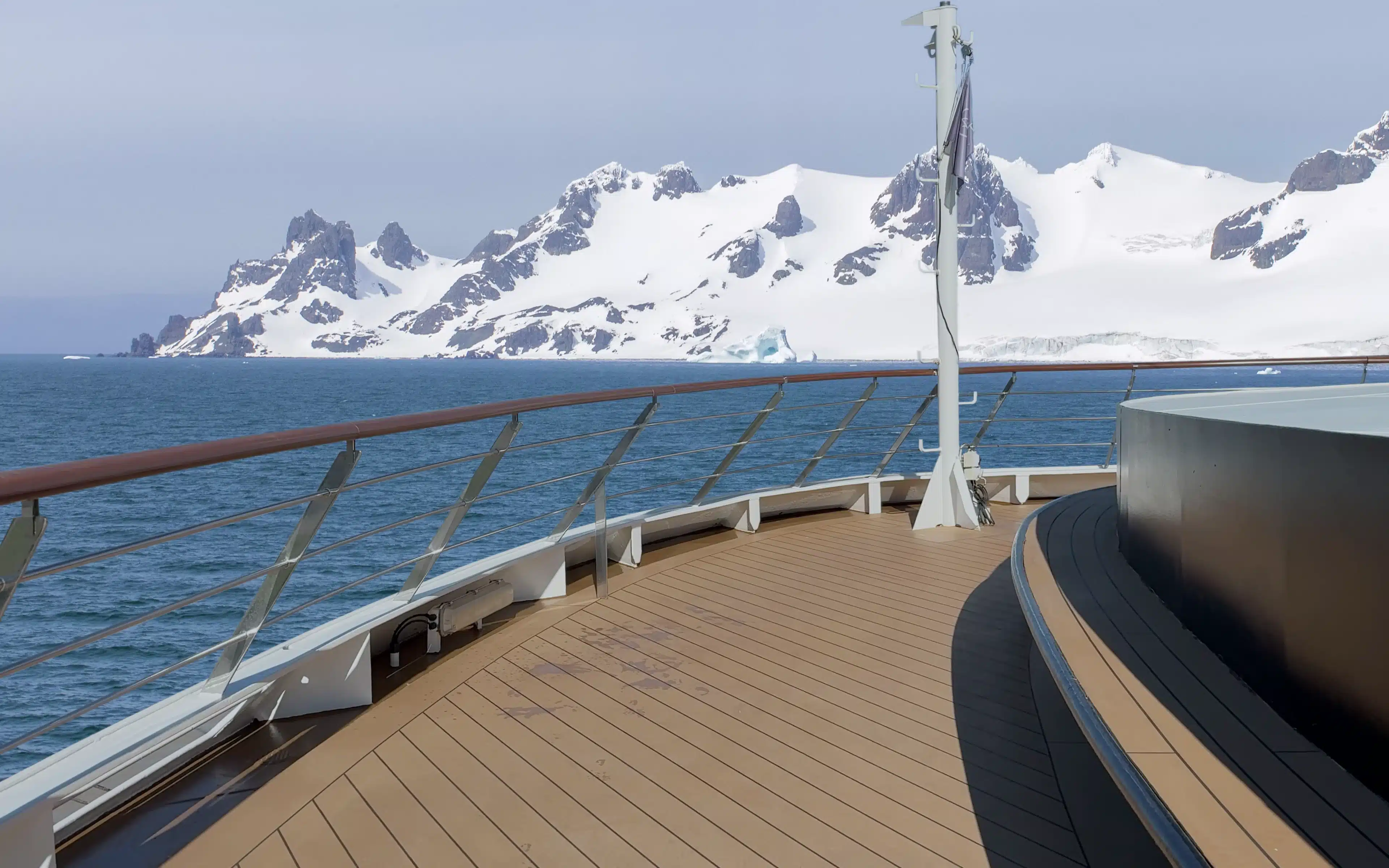 World Voyager forward observation deck with teak decking and polished steel railings, looking out across deep blue polar waters toward dramatic snow-capped Antarctic mountains and glaciers under a pale winter sky