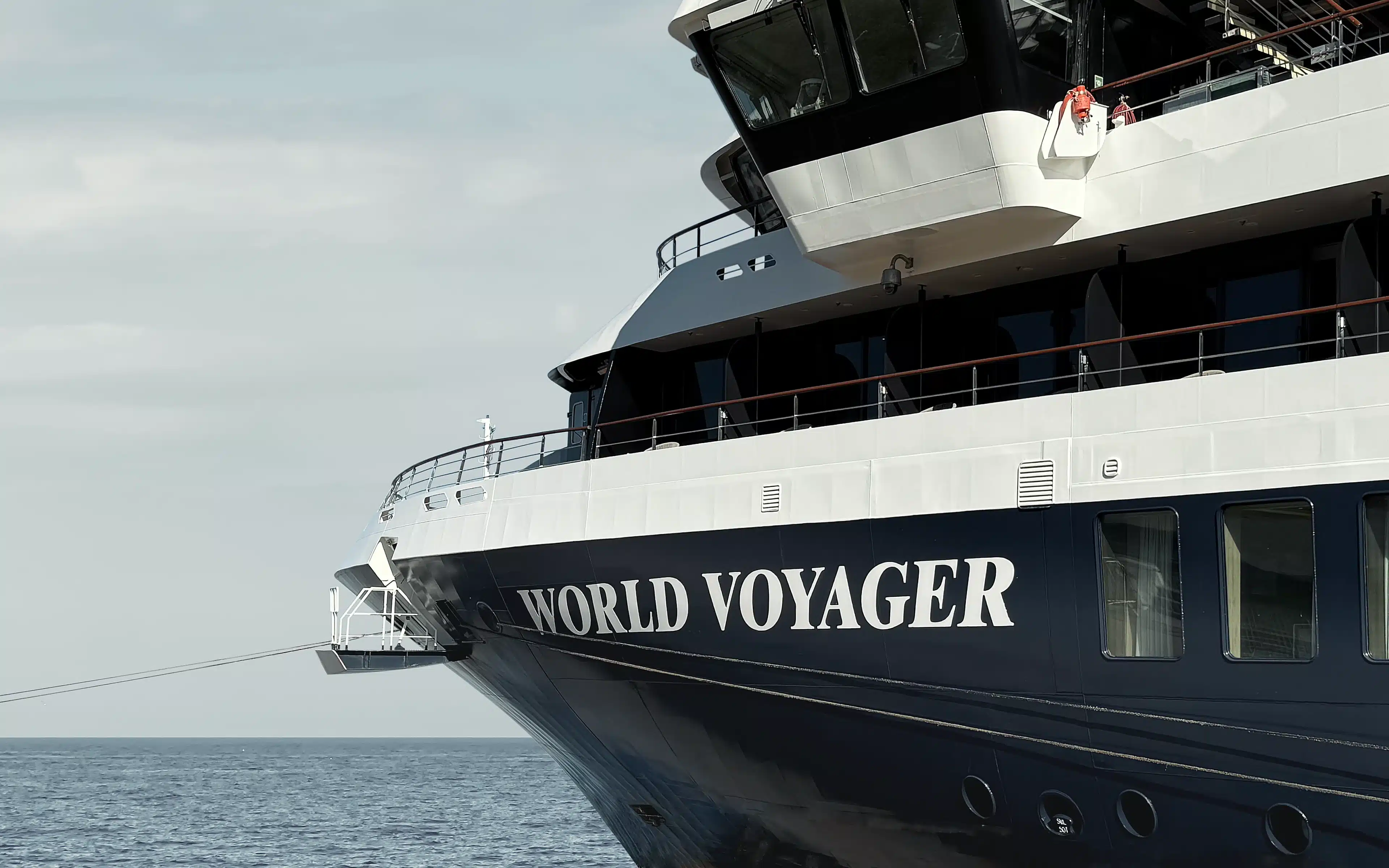 The bow of Atlas Ocean Voyages' World Voyager at anchor, showing the vessel's deep navy hull with name in white lettering, white superstructure, and forward observation deck against a calm grey-blue Atlantic horizon