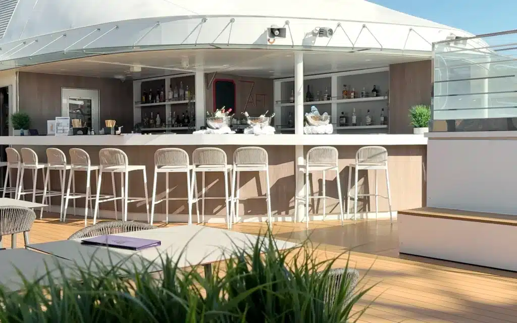 World Voyager 7AFT Pool Bar from a wider angle showing the full bar counter, white rope stools, a stocked spirit shelf, champagne in ice buckets, and a clear blue sky beyond the Dome structure