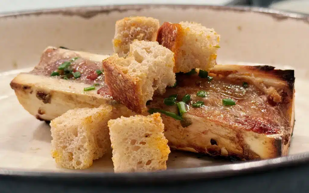 AFT Grill charcoal-grilled beef bone marrow halves with golden croutons and chives, served in a shallow speckled bowl