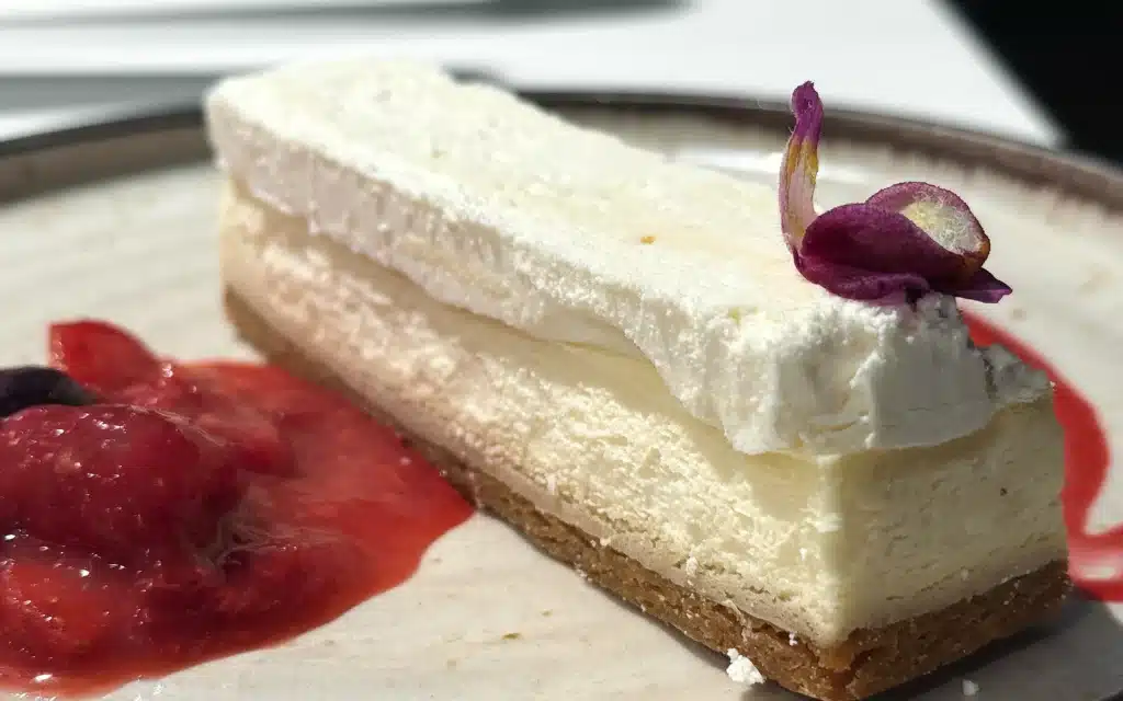 7AFT Grill cheesecake slice with a masterfully constructed shortcrust pastry base, light cream cheese filling, edible purple flower, and a berry compote on a speckled plate