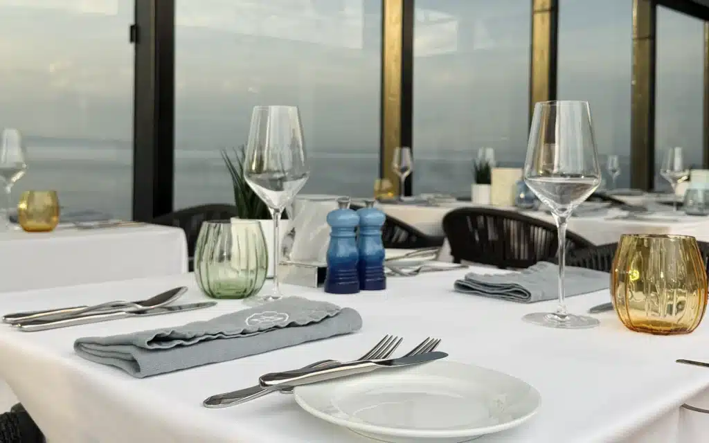 7AFT Grill restaurant interior close-up showing white linen place settings with silver cutlery, green and amber glass votives, blue ceramic salt and pepper grinders, and coastal ocean views through floor-to-ceiling windows