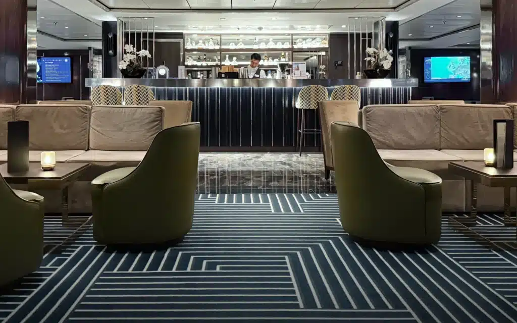 World Voyager Atlas Lounge interior with olive green curved armchairs, taupe sofas, and a bartender at work behind a glowing bar, geometric navy carpet, and soft ambient lighting