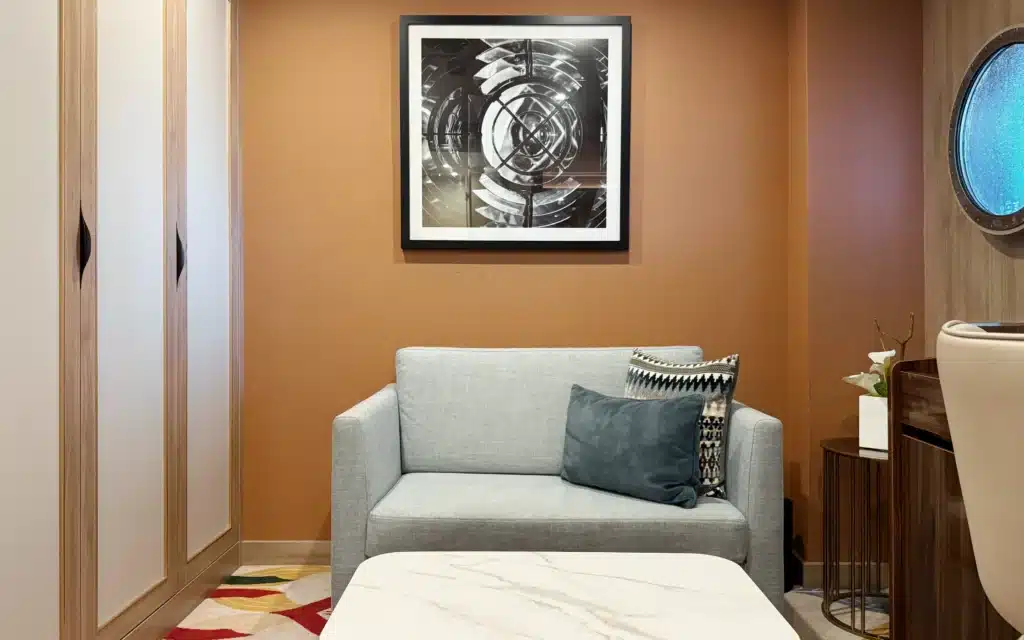 Separate living area of a World Voyager Deluxe Veranda Suite with a grey linen sofa, marble-topped coffee table, botanical patterned carpet, and a framed nautical black-and-white artwork above a porthole window
