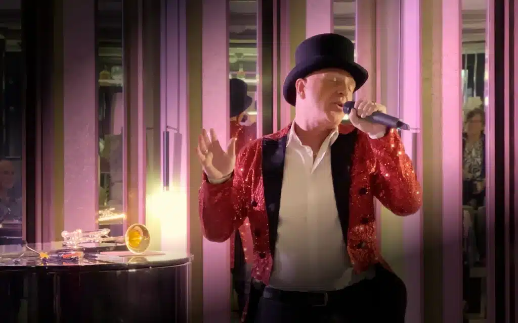 World Voyager onboard entertainment performance showing a male vocalist in a sequined red jacket and black top hat singing into a microphone under warm pink stage lighting, with a trumpet visible on a side table behind him