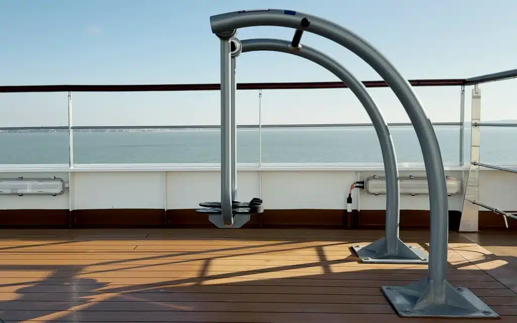 Open-deck exercise station aboard World Voyager featuring a stainless steel arc-shaped pull-up and stretching frame bolted to teak decking, with a calm Atlantic Ocean and distant coastline visible beyond the railings