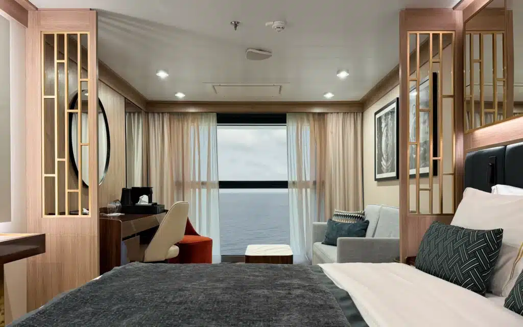 World Voyager Horizon Suite alternative room view showing the king bed, sitting area, floor-to-ceiling retractable window open to Atlantic Ocean views, warm oak paneling, gold lattice screens, and framed nautical artwork