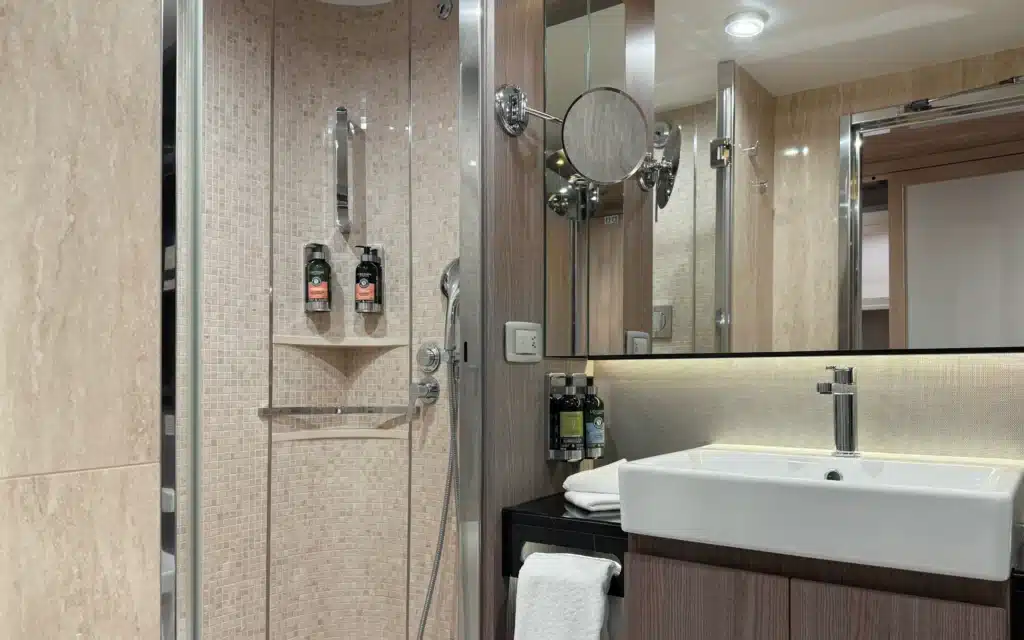World Voyager Horizon Suite bathroom featuring a curved glass-enclosed mosaic-tile shower with rain head and handheld fitting, L'Occitane amenities, vessel sink on a dark vanity, and a wall-mounted magnifying mirror