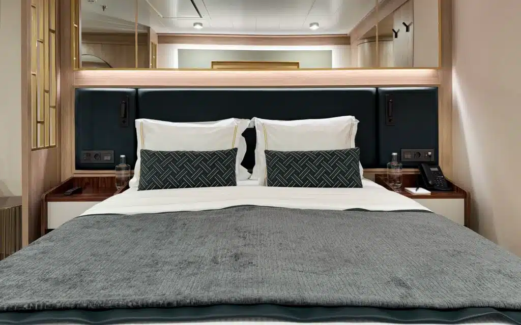 World Voyager Horizon Suite king bed with dark teal upholstered headboard, integrated ambient lighting, herringbone accent cushions, gold-trimmed white pillows, twin bedside tables, and branded still water bottles