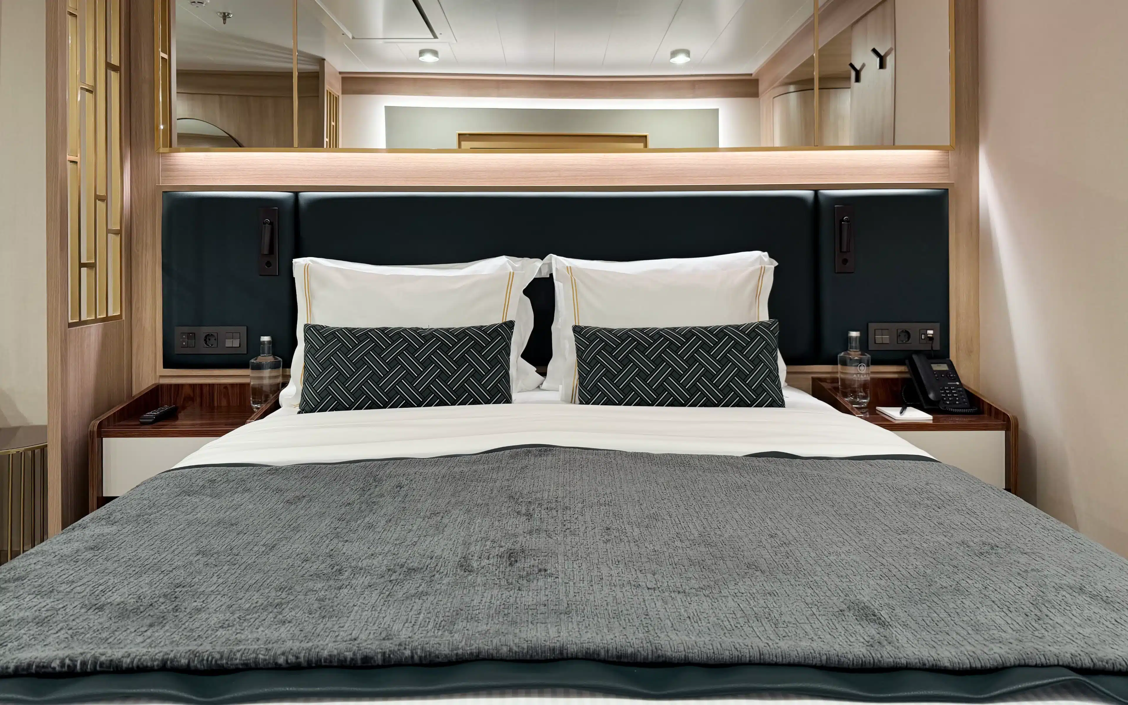 World Voyager Horizon Suite king bed with dark teal upholstered headboard, integrated ambient lighting, herringbone accent cushions, gold-trimmed white pillows, twin bedside tables, and branded still water bottles