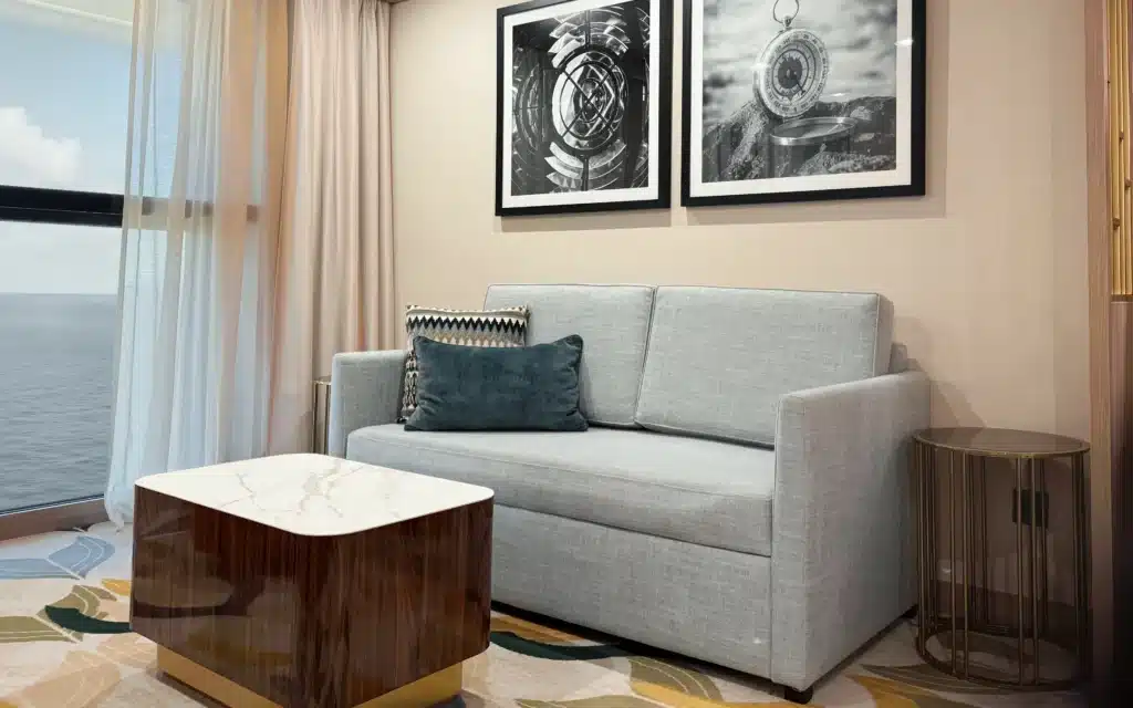 orld Voyager Horizon Suite sitting area with a grey linen two-seat sofa, teal velvet accent cushion, marble-topped coffee table, gold side table, two framed black-and-white nautical artworks, and Atlantic Ocean views through floor-to-ceiling curtains