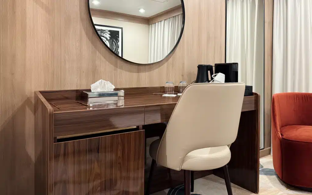 World Voyager Horizon Suite writing desk with a dark walnut finish, cream leather chair, round black-framed mirror, tissue box, and Nespresso station visible beside a glimpse of the sitting area's red accent chair