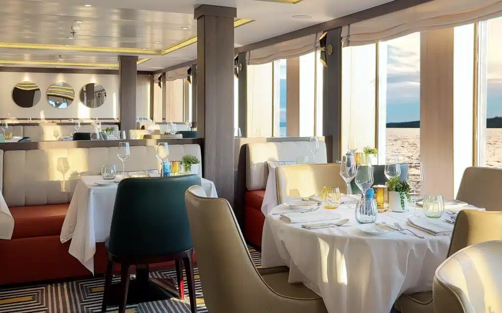 World Voyager Madeira Restaurant interior at golden hour showing white-linen round tables with crystal wine glasses, coloured glass votives, succulent centerpieces, teal and cream leather dining chairs, terracotta red banquette seating, round porthole mirrors, and floor-to-ceiling windows framing a luminous Atlantic Ocean sunset