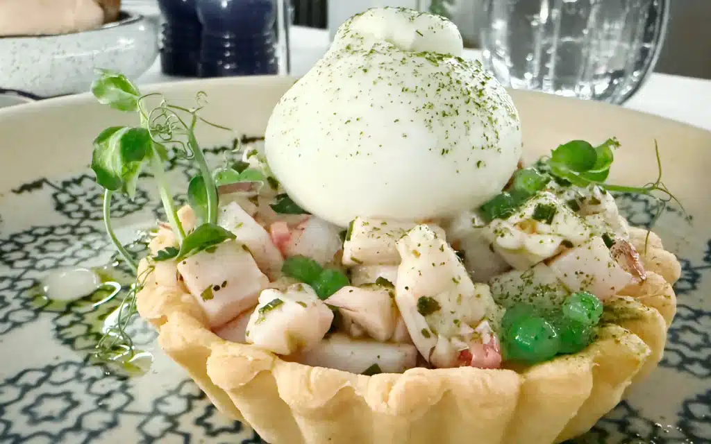 Madeira Restaurant seabass tartare served in a pastry tartlet shell, topped with a burratina sphere dusted with green herb powder, surrounded by pea pearls and microgreens on a patterned plate