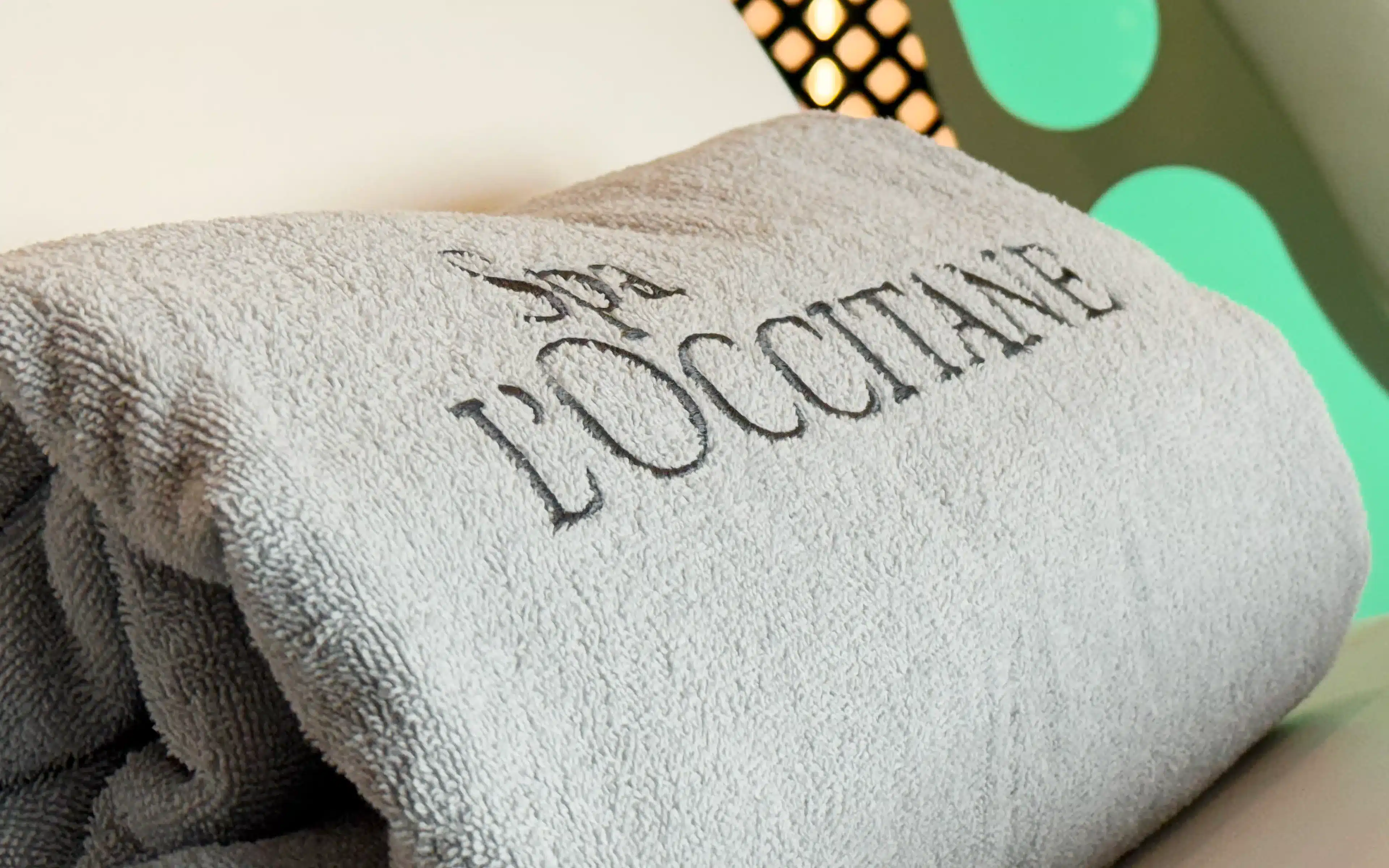 Close-up of a folded grey L'Occitane branded towel in the SeaSpa relaxation room aboard World Voyager, with colourful geometric wall art visible in the background