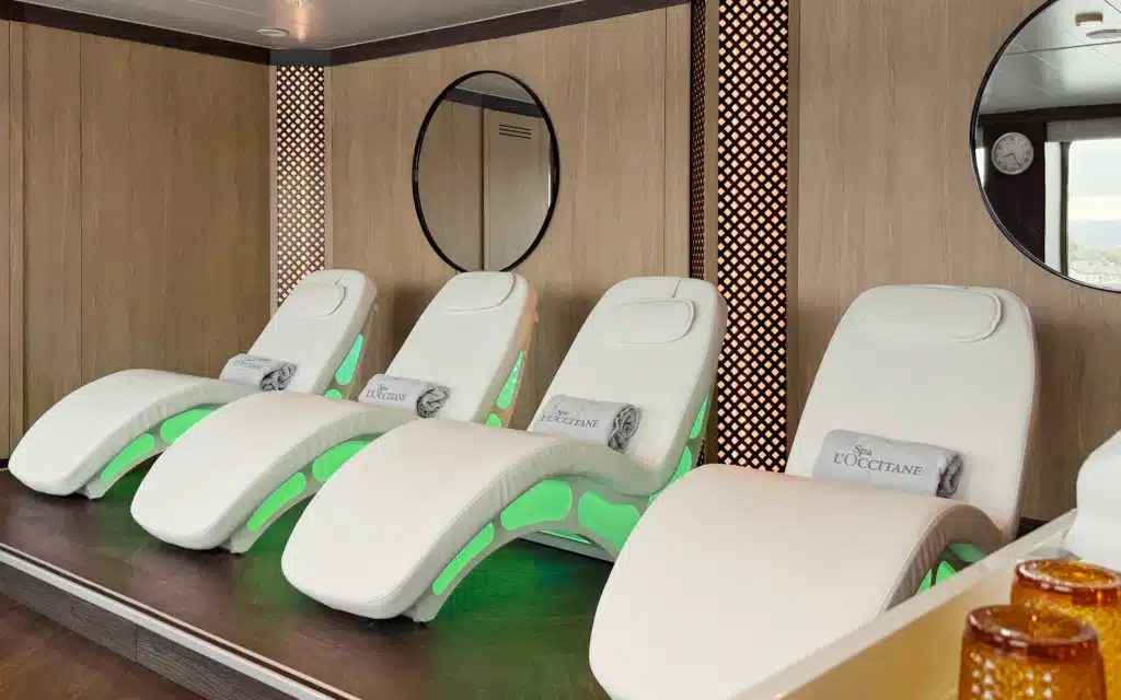 SeaSpa by L'Occitane relaxation room aboard World Voyager featuring a row of illuminated white heated loungers with green lighting accents, L'Occitane branded towels, and amber glass vessels in the foreground