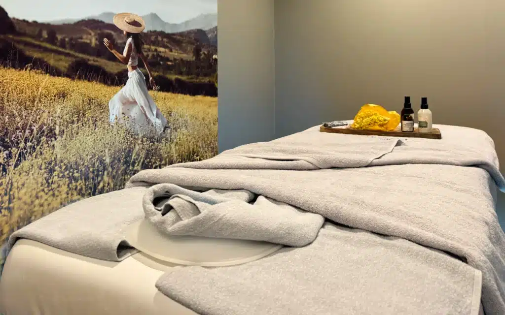 SeaSpa by L'Occitane treatment room aboard World Voyager with a prepared massage table draped in grey towels, a wooden tray of treatment products, and an illustrated floral mural backdrop