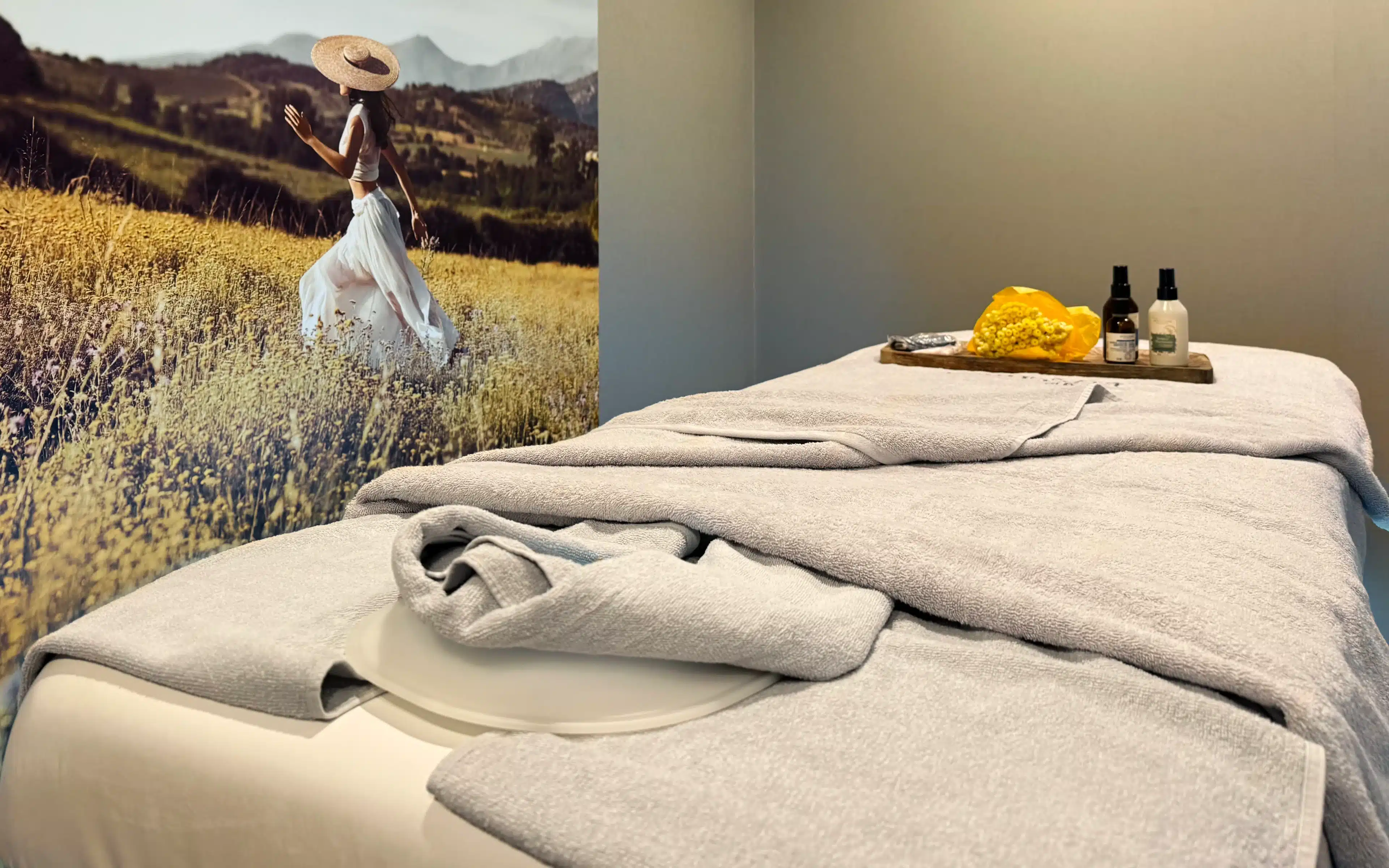 SeaSpa by L'Occitane treatment room aboard World Voyager with a prepared massage table draped in grey towels, a wooden tray of treatment products, and an illustrated floral mural backdrop