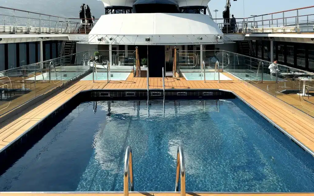 World Voyager outdoor heated swimming pool viewed from the entry end in port with teak decking, glass railings, whirlpool tubs on an elevated platform, and a mountainous coastline visible beyond