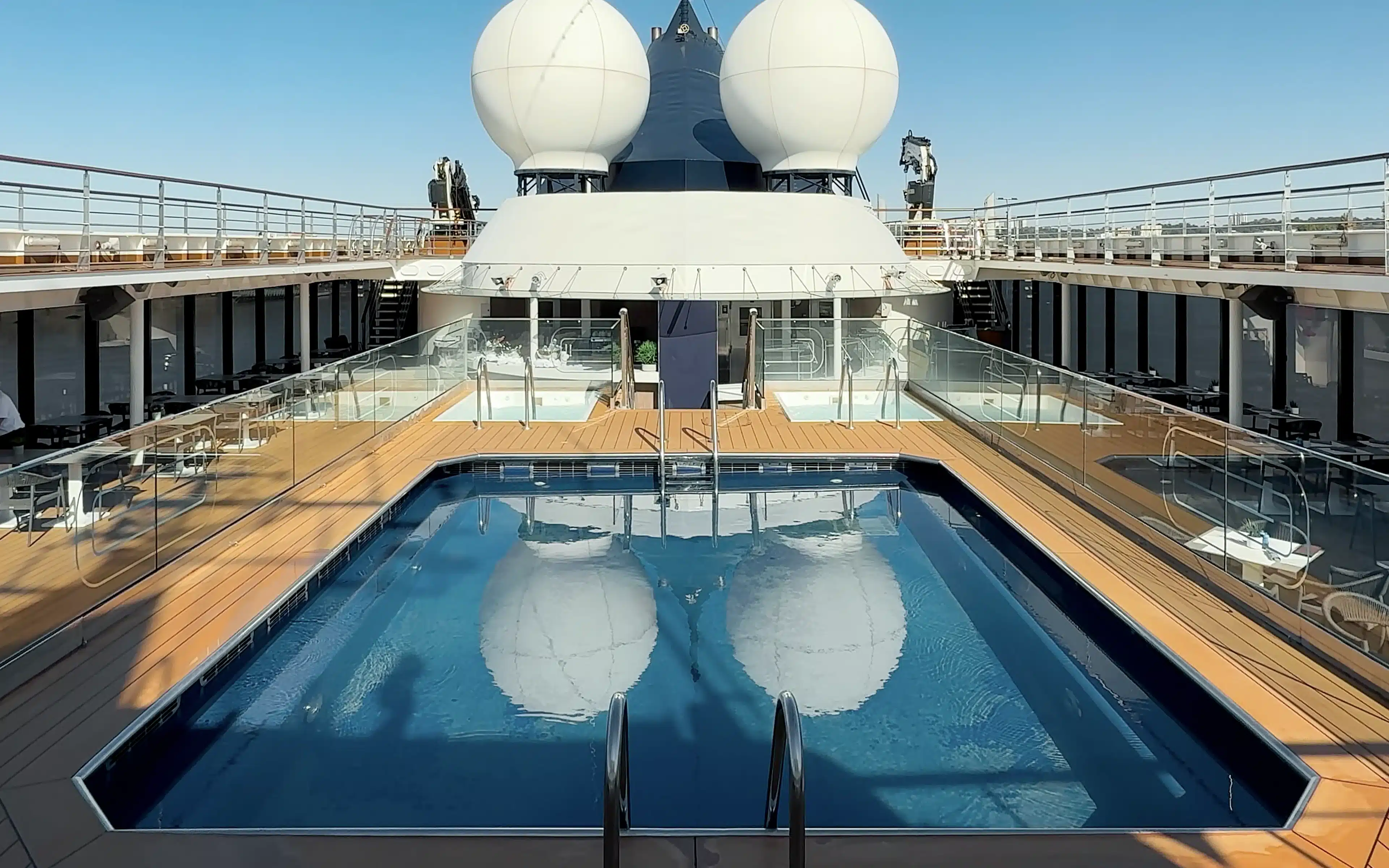 World Voyager outdoor heated swimming pool viewed from the entry steps on a clear blue-sky day, with teak pool decking, glass railings, two whirlpool tubs visible beyond, and the ship's white superstructure and twin radar domes overhead